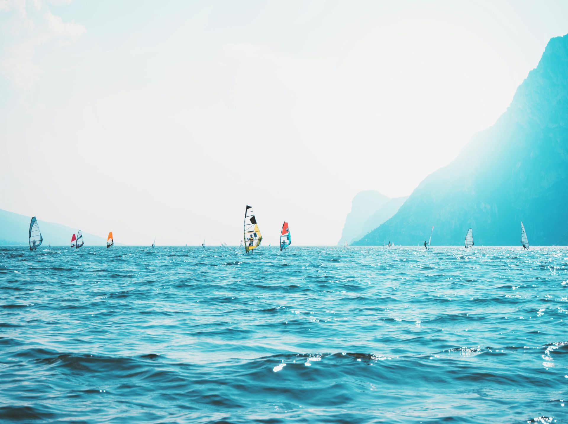 The image showcases people windsurfing on a sunny day with mountains in the background and a vibrant blue sea in the foreground.