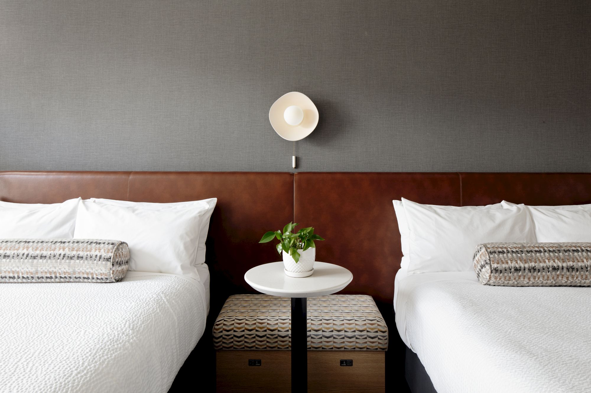 A modern hotel room with two beds, white pillows, a small side table, a potted plant, and a ceiling lamp above.