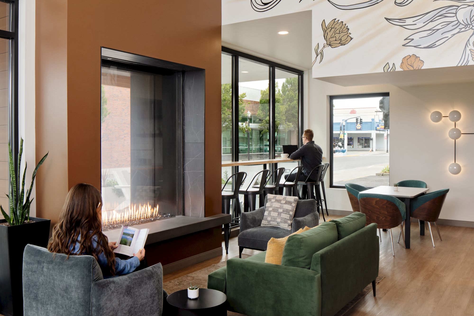 A modern lounge with a glowing fireplace, a mural wall, people relaxing and reading, and large windows looking out to the street.