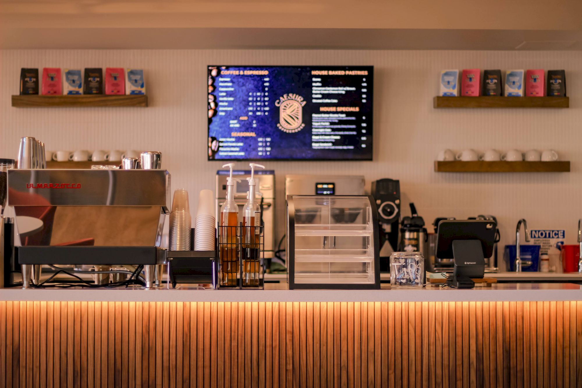 A cafe counter with a menu screen, espresso machine, mugs, and shelves of colorful cups, warm lighting, and a striped front panel.
