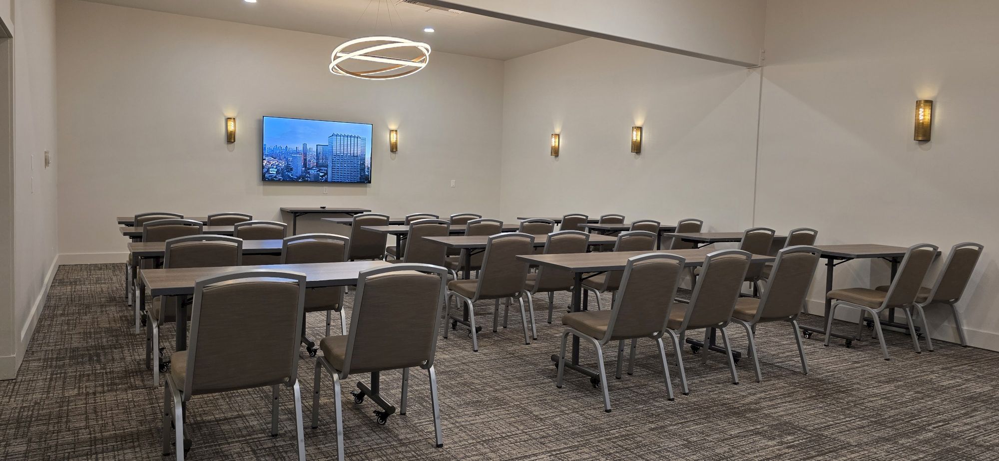 A conference room with rows of gray chairs facing a wall-mounted screen, elegant circular light fixture, and wall sconces in a neutral, professional setup.