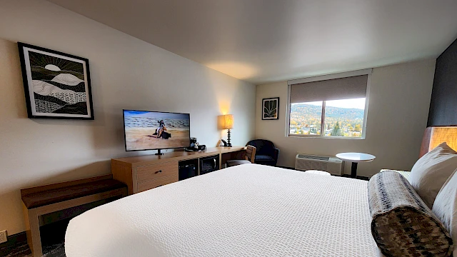 A hotel room with a large white bed, a desk and TV, a small seating area by a window, framed art on the walls, and soft lighting.
