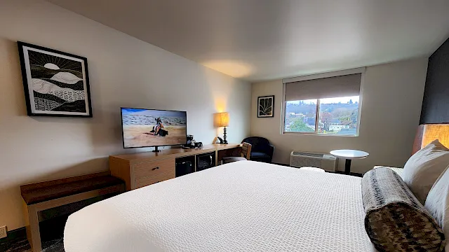 A hotel room with a large bed, desk area, flat-screen TV, a bench, and a window view; cozy lighting and a round table by the window.