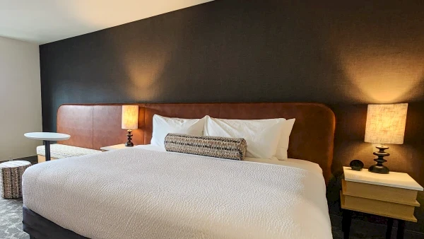 A luxurious hotel room with a large bed, cozy two lamps, a dark accent wall, and modern bedside tables and stools, inviting and tidy.