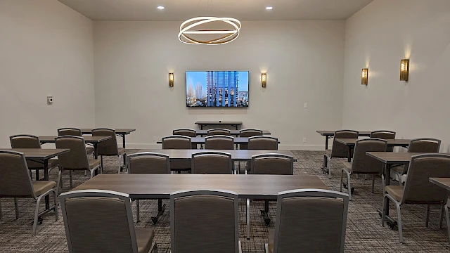 A conference room setup with multiple rectangular tables and chairs facing a wall-mounted screen displaying a cityscape, under modern pendant lights.