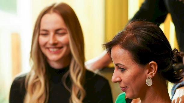 Two stylish women sit at a table, one in a green sweater signing papers while another smiles in the background, creating a professional vibe.