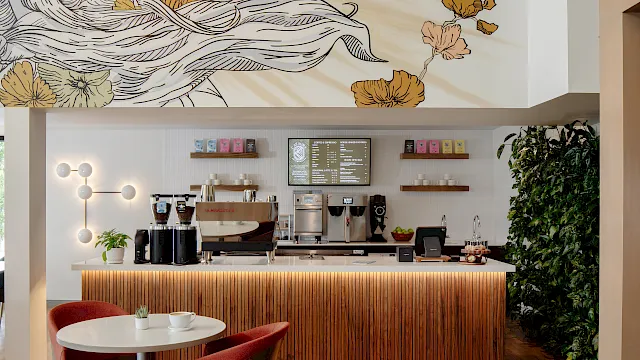 A modern coffee shop counter with espresso machines, shelves of coffee and pastries, wall art of flowers, and seating in front.