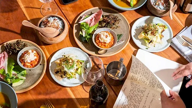 A top-down dinner scene with plates of food, drinks, and a person holding a menu or paper, surrounded by a warm wooden table.