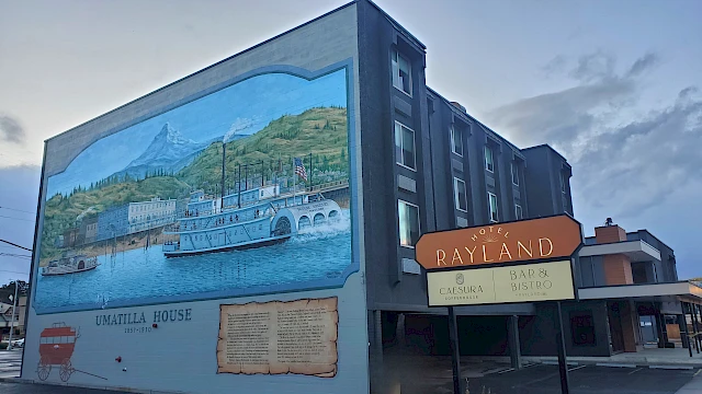 A building with a large mural of a scenic harbor and boat, labeled &ldquo;Umatilla House,&rdquo; beside a street sign for Rayland Inn, under a cloudy sky.