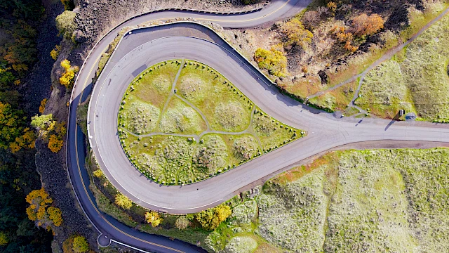 A winding hillside road loops around a grassy, fenced patch with small circular plots, surrounded by autumn trees and steep slopes.