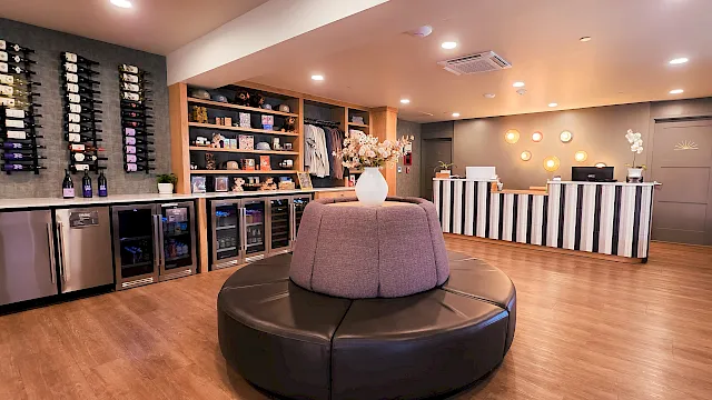 A modern lounge or hotel lobby with a circular cushioned seating area in the center, a reception desk to the right, and drinks or small shelves along the left wall under warm interior lighting.