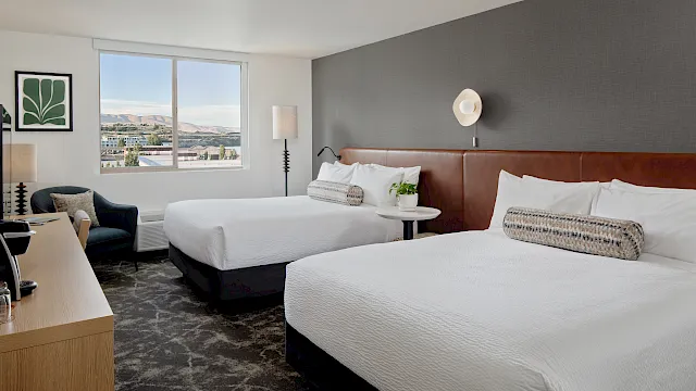 A modern hotel room with two white beds, bedside lamps, and a large window showing a scenic mountain view.