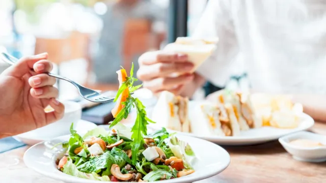 Two people share a fresh salad with greens and nuts at a table, while one holds a sandwich in a cozy cafe.