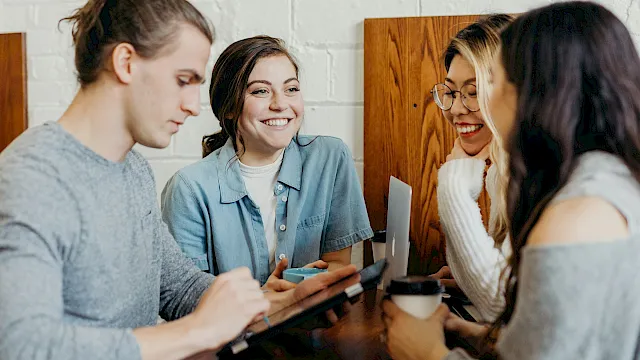 Four friends sit around a table chatting and smiling, one holding a phone while another writes on a tablet, coffee cups nearby.