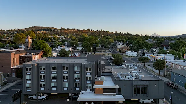A suburban town skyline with streets, parked cars, buildings, a clock-tower, and green hills under a clear blue sky.