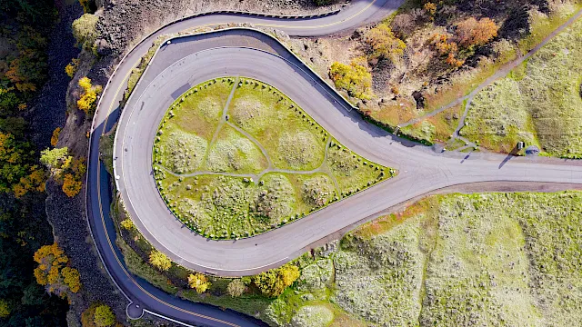 Aerial view of a winding road forming a loop through grassy, hilly terrain with autumn trees, sharp hairpin turns, and a gate-like curve.