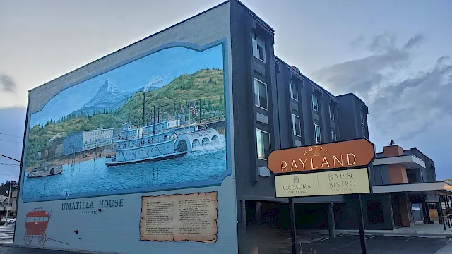 A mural on the side of a building depicts a river town with boats, hills, and a large &ldquo;Umzila House&rdquo; plaque; a sign reads &ldquo;Rayland.&rdquo;