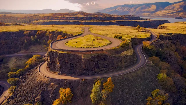 An aerial view of a ancient circular fortress on a hill, winding roads spiraling around rugged terrain with autumn trees under a wide sky.