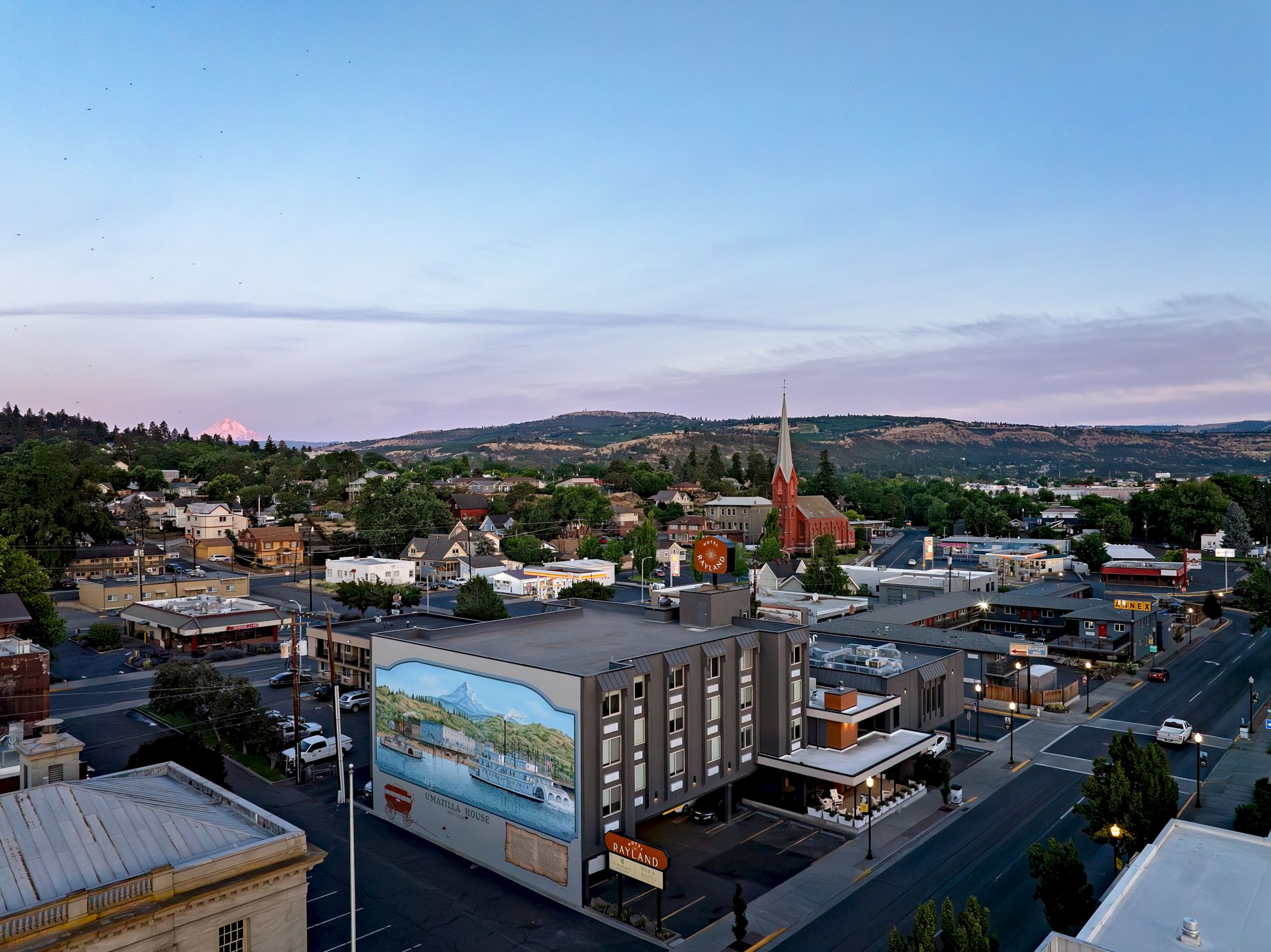 A small town street scene with a church steeple, hotels, and distant hills under a blue evening sky.