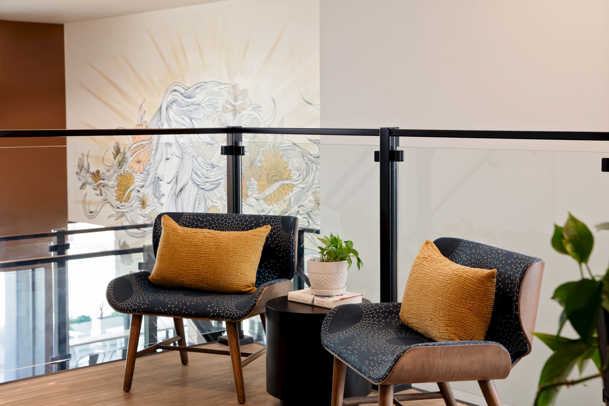 A modern lounge with dark chairs and gold cushions, a small table with a book and plant, and a decorative wall mural behind.