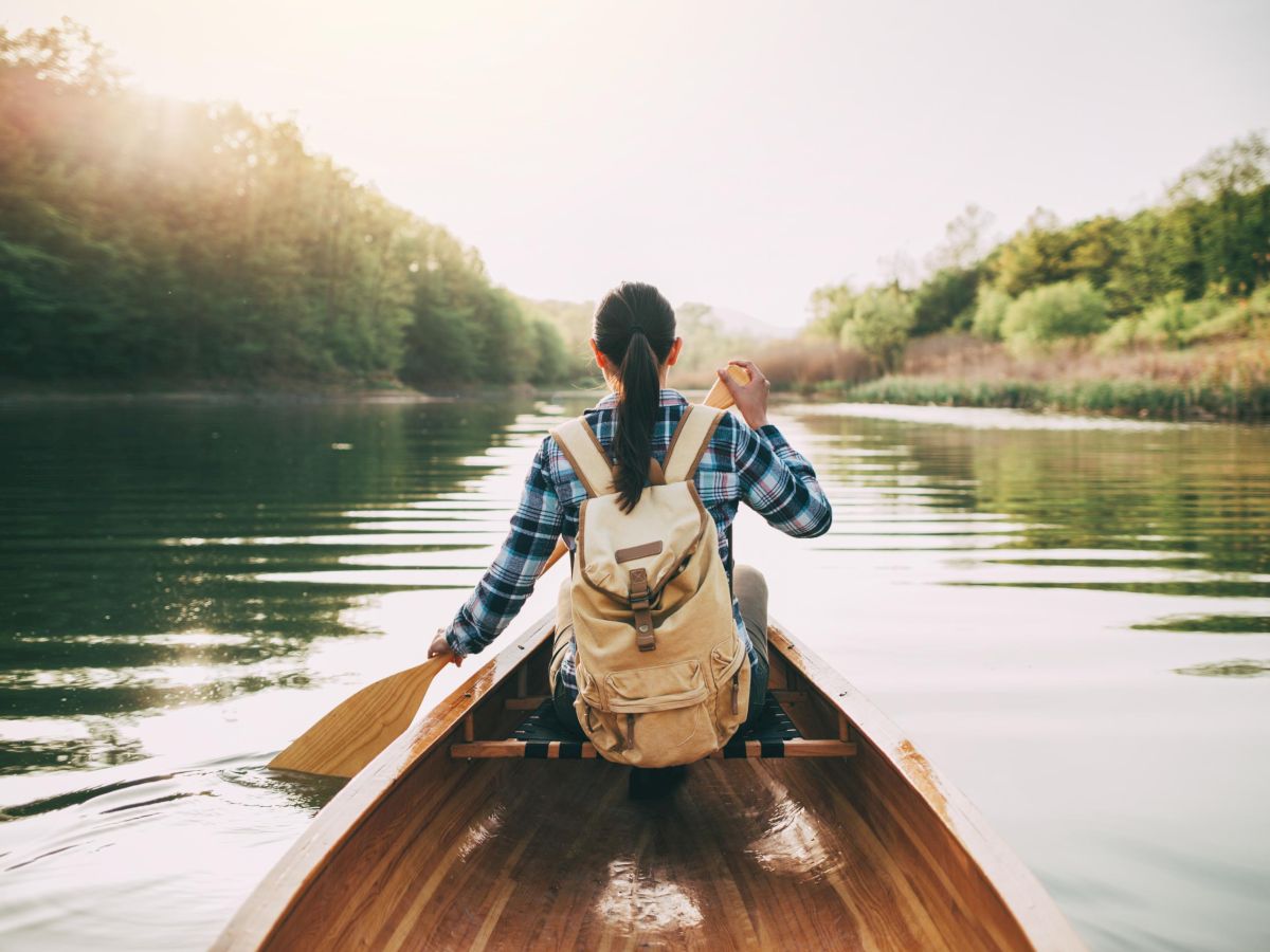 A traveler in a canoe paddles down a calm river at sunrise, wearing a plaid shirt and backpack.