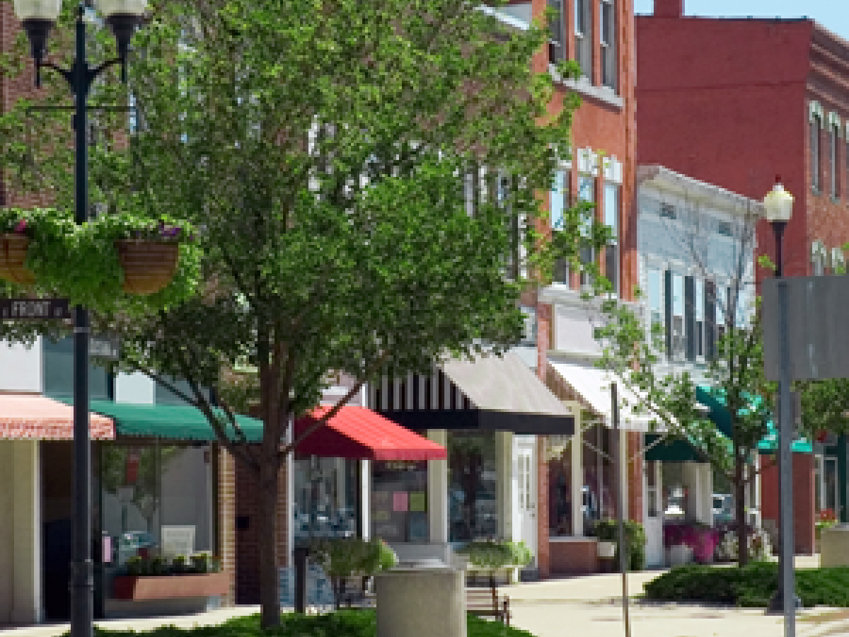 A sunny downtown street with green trees, lampposts, and colorful storefront buildings lining the sidewalk.